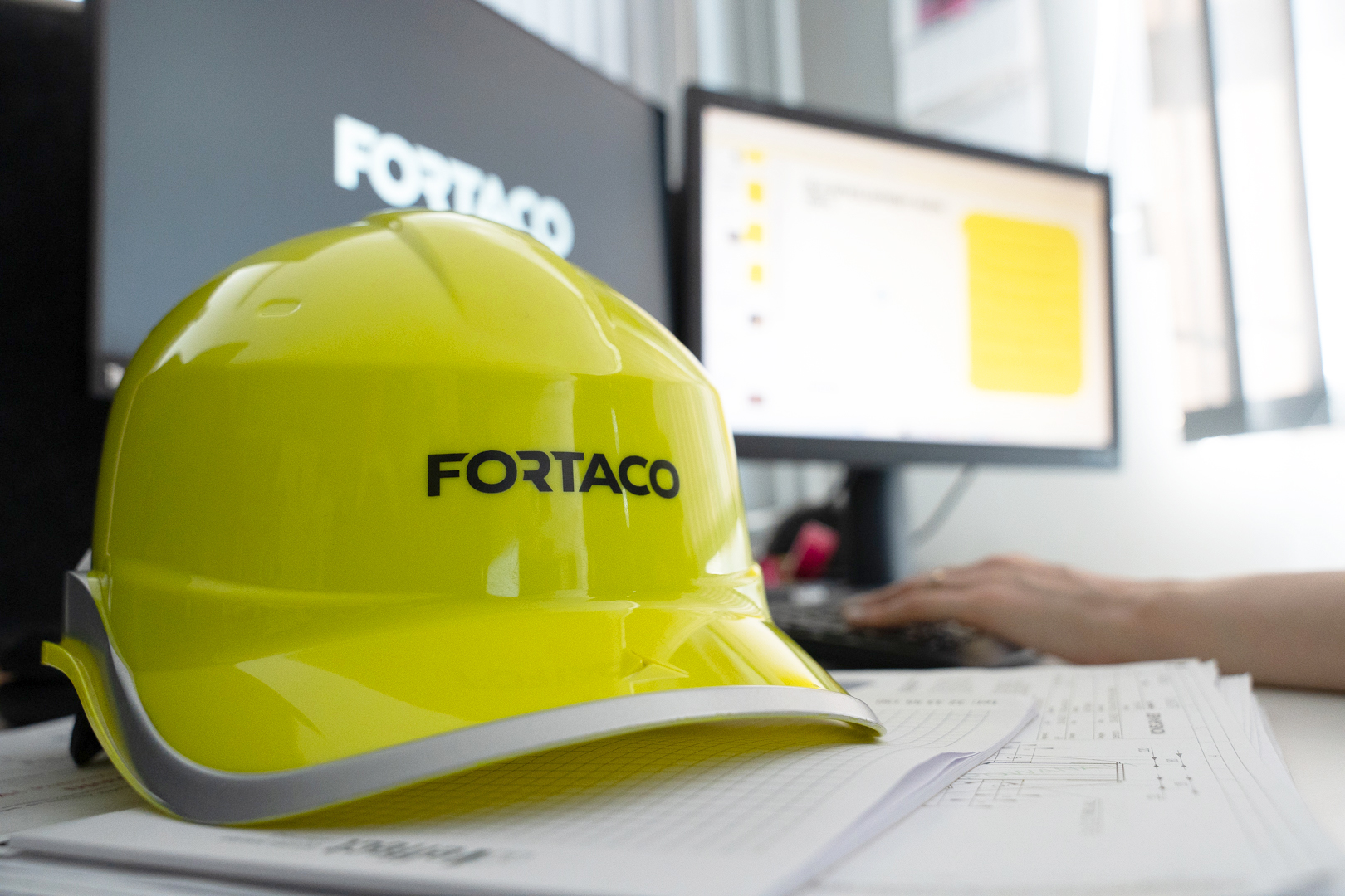 A yellow hard hat with "FORTACO" written on it rests on top of technical drawings, in front of a person working at a desk with computer screens in the background.