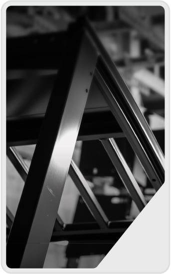 Black and white photo showing a close-up of a metal framework or truss with diagonal and horizontal beams, creating geometric shapes and patterns. Background is blurred, emphasizing the structure in the foreground.