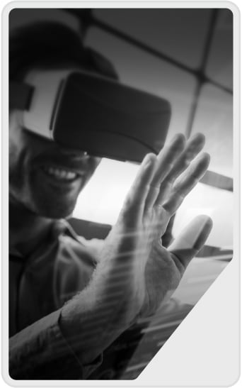 A person wearing virtual reality goggles smiles and reaches out with one hand, interacting with an unseen interface. The image is in black and white, with a futuristic, immersive atmosphere.