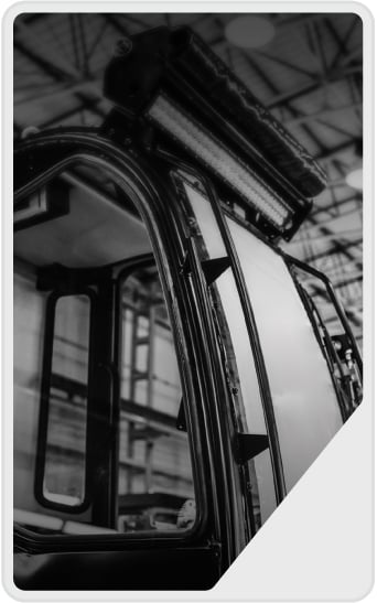 Black and white photo of a vehicle cab, possibly a construction or industrial machine, with a curved window and mounted lights on the roof, inside a large building or warehouse.