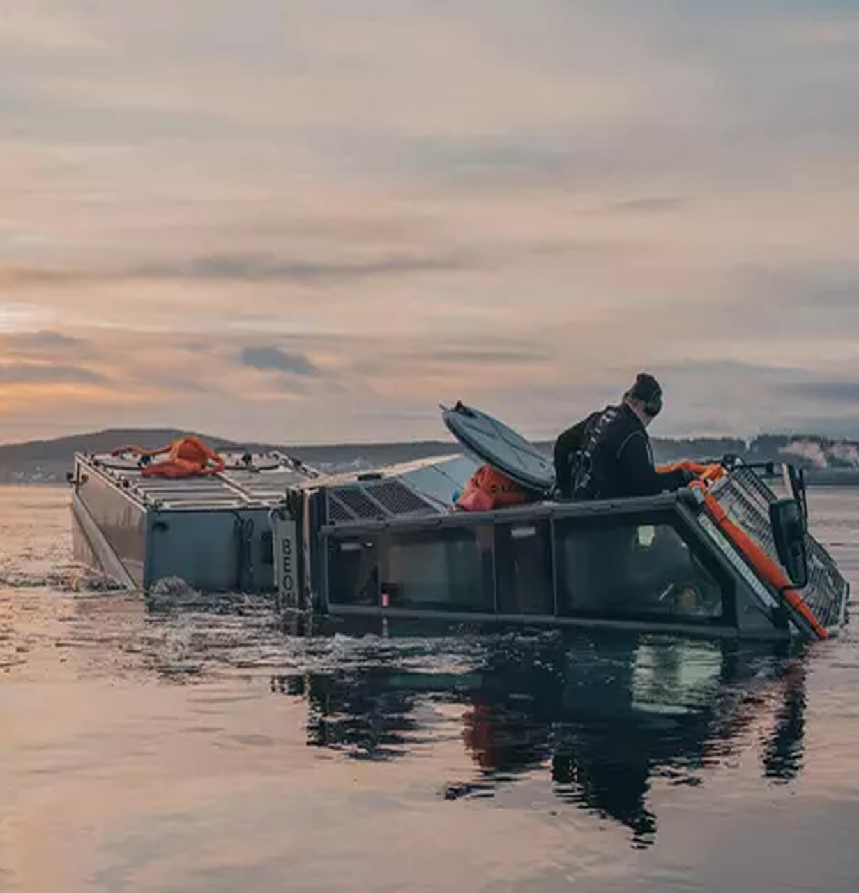A person stands on top of a partially submerged vehicle in calm water at sunset, with distant hills and buildings visible in the background.