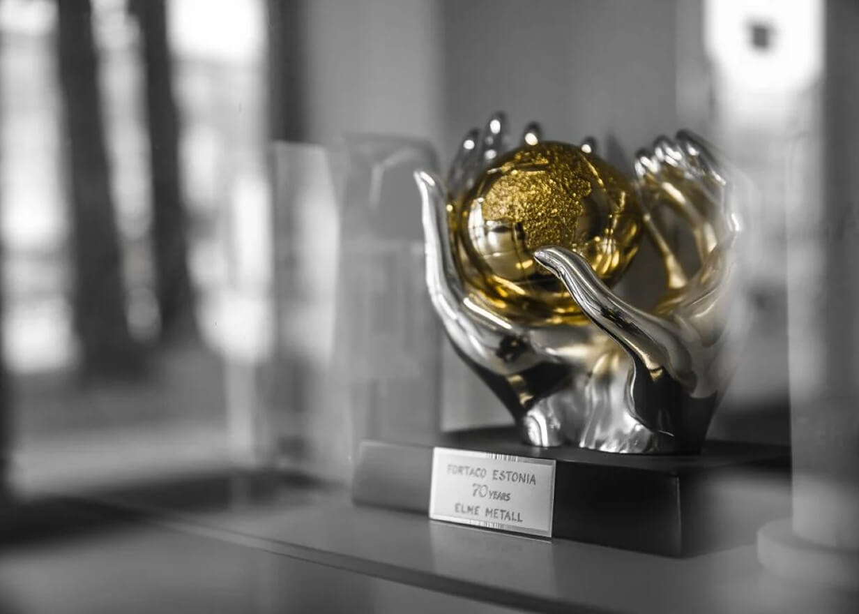 A shiny silver trophy shaped like hands holding a golden globe sits on a reflective surface. The base has a plaque that reads, "Fortaco Estonia 70 Years Elme Metall." The background is blurred.
