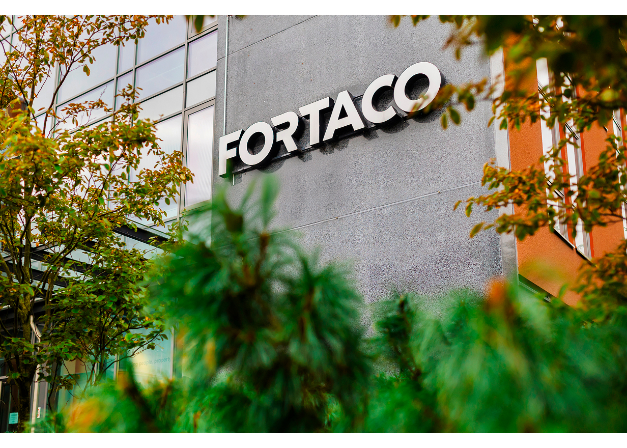 A building exterior with the large, modern white "FORTACO" sign mounted on a gray wall, surrounded by green leafy plants and trees.