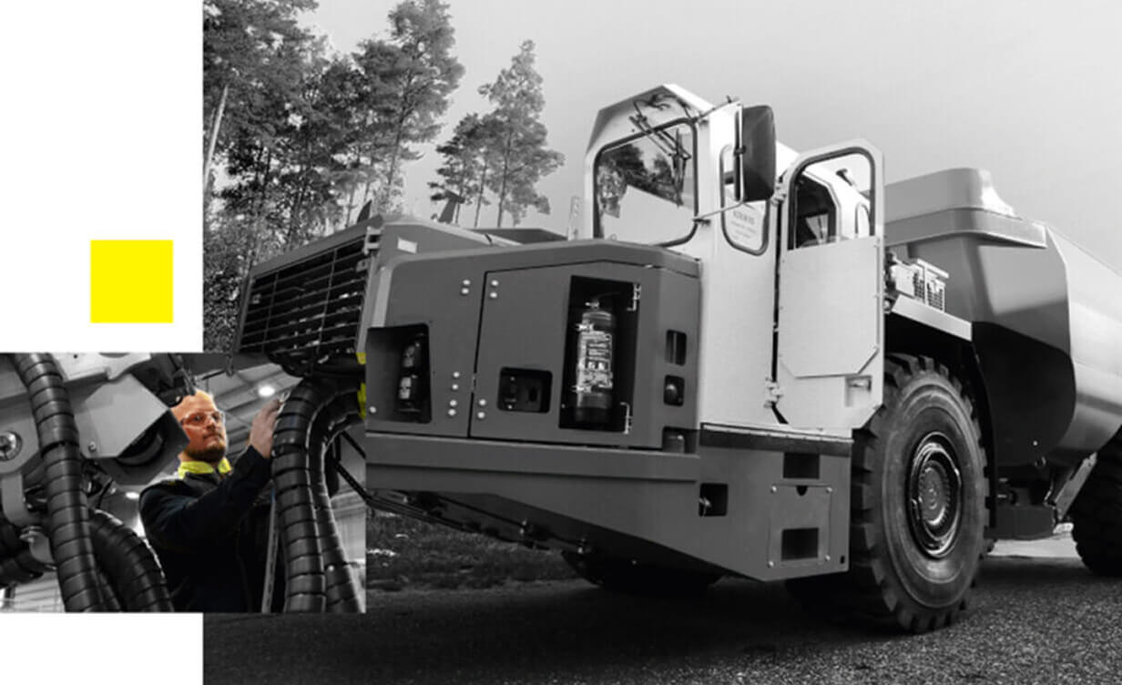 A person wearing safety glasses inspects or repairs a large industrial dump truck on a road, with trees in the background. The image is mostly in grayscale with a few yellow accents.