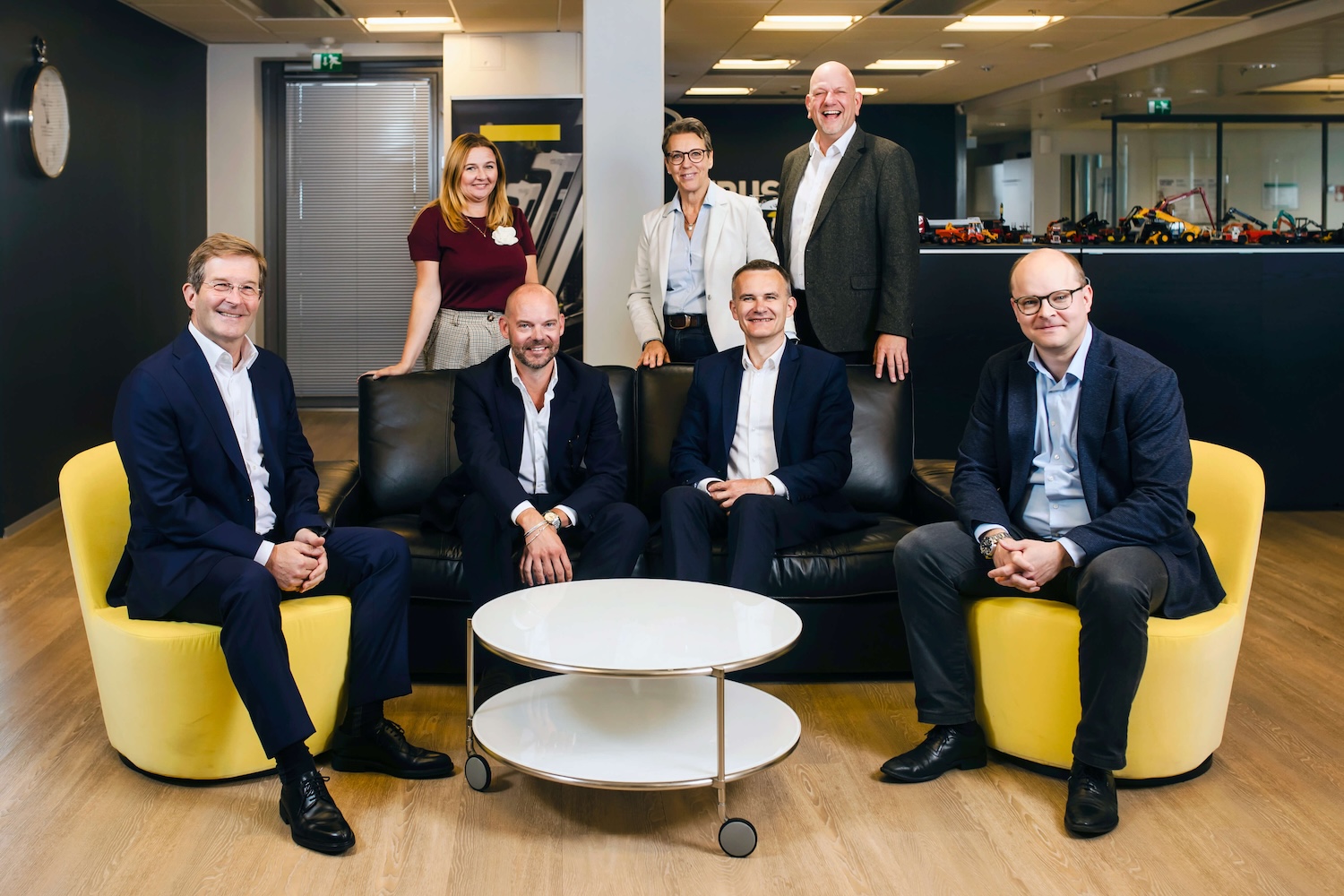 Seven professionally dressed people, four seated and three standing, pose and smile in a modern office with yellow chairs, a black couch, and a round white coffee table.