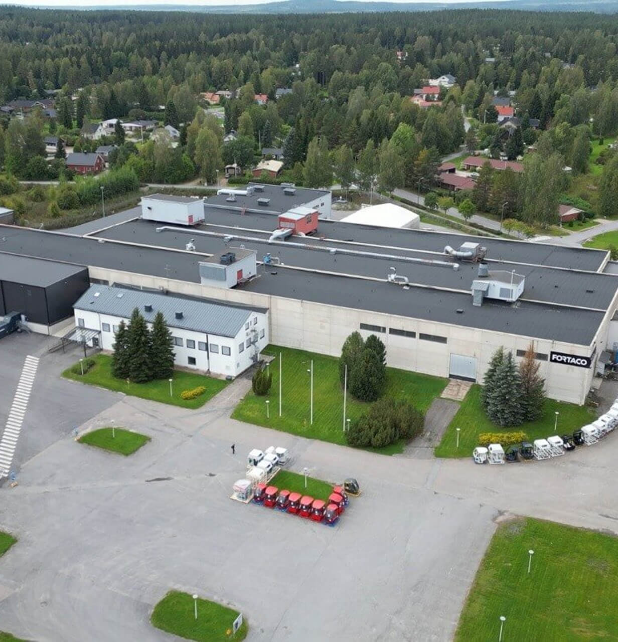 Aerial view of a large industrial building surrounded by trees, with several red vehicles parked in a row outside and a few white vans nearby. Residential houses and dense forest are visible in the background.