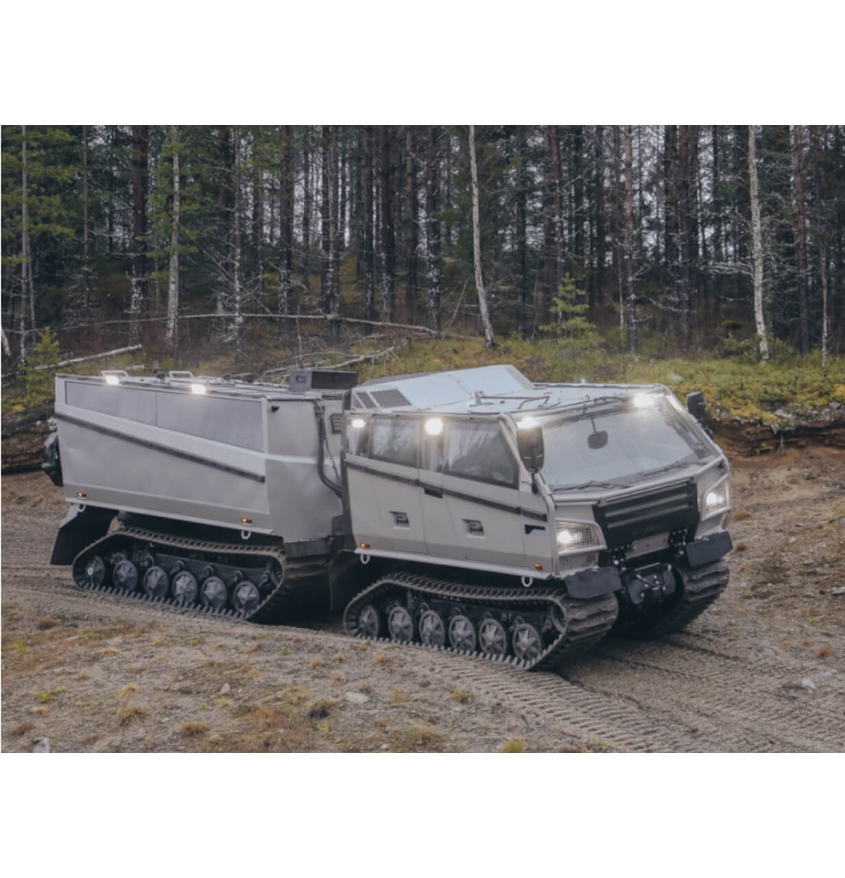 A large, gray, all-terrain tracked vehicle with two connected segments drives through a forested area on a dirt path, surrounded by trees and greenery.