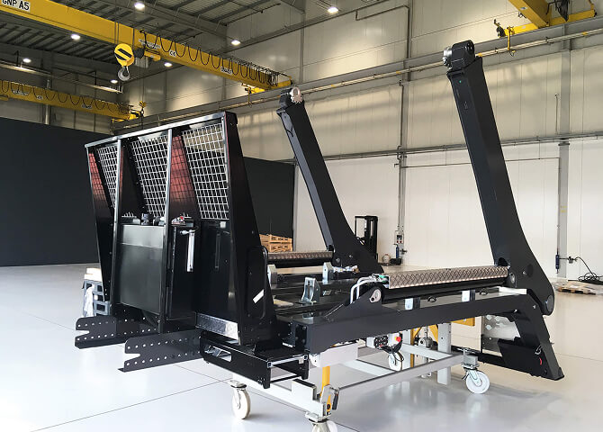 A large industrial machine with metal arms and mesh panels stands in a spacious, well-lit warehouse with overhead cranes and high ceilings.