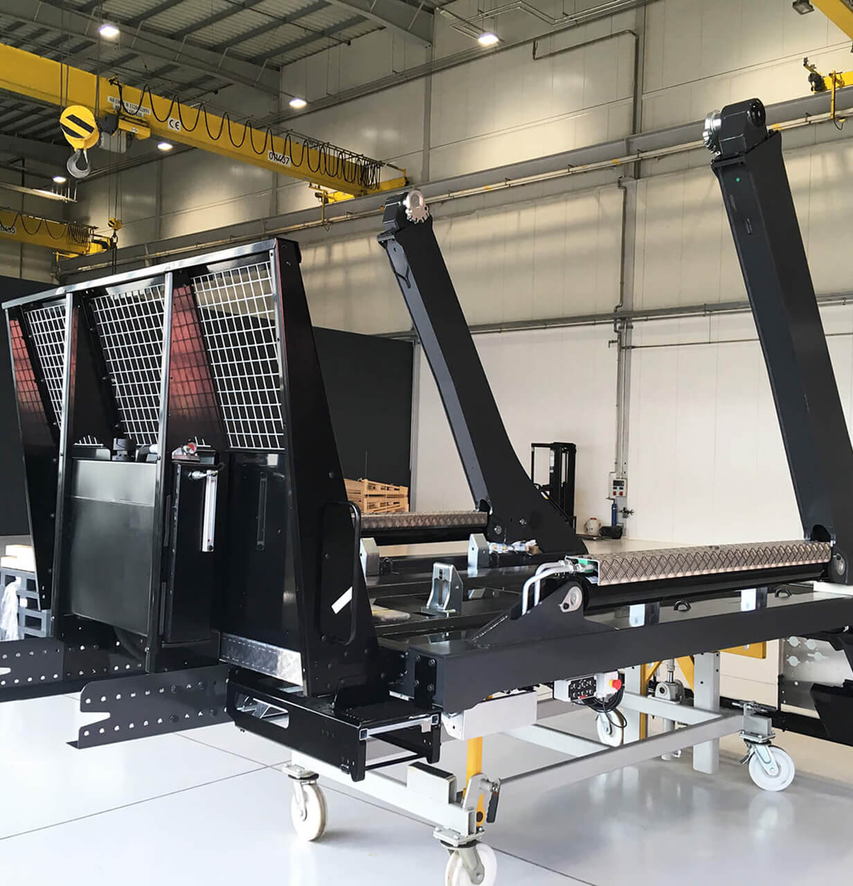 A large industrial conveyor system with black metal frames and rollers is set inside a spacious, well-lit factory with yellow overhead cranes and white walls and floors.