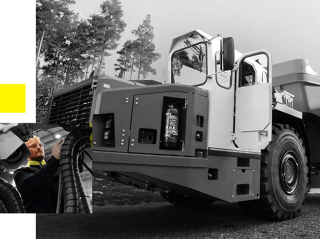 A worker inspects the large front tire of an industrial dump truck parked outdoors, with trees visible in the background. The image is mostly black and white, except for some yellow highlights.