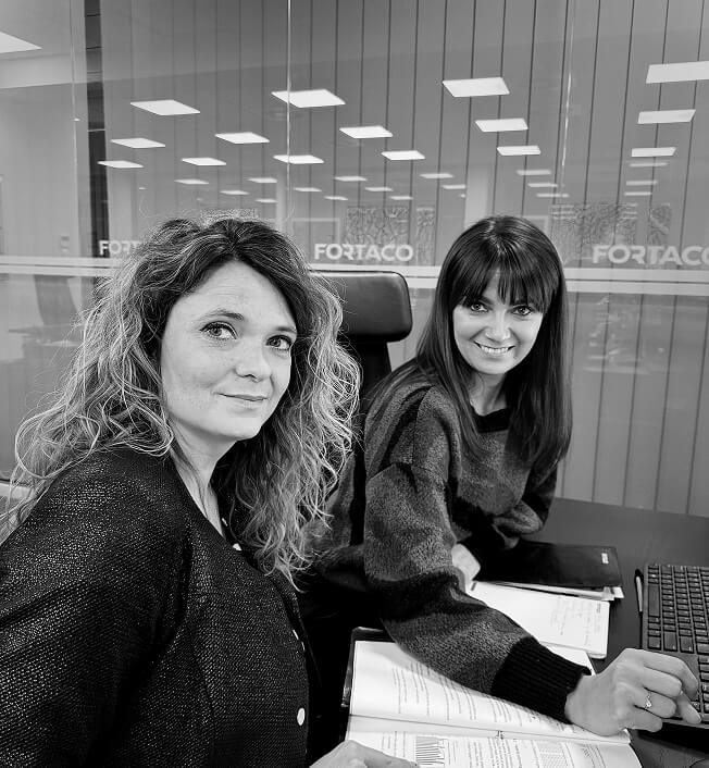 Two women sit at a desk in an office, smiling at the camera. They have open documents in front of them and a keyboard nearby. The background shows glass walls with the word "FORTACO" visible.