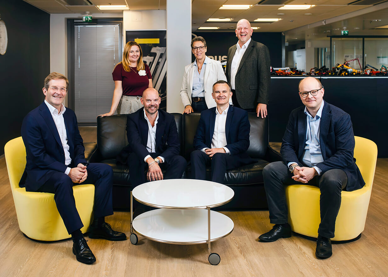 Seven professionally dressed people, four men seated on a black sofa and three people standing behind, pose and smile in a modern office space with yellow chairs and a white round table in front.