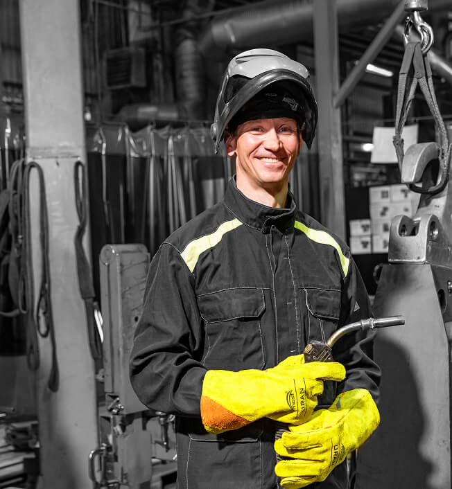 A smiling worker in a protective helmet, safety glasses, black coveralls with yellow accents, and yellow gloves stands in an industrial setting holding a welding torch.