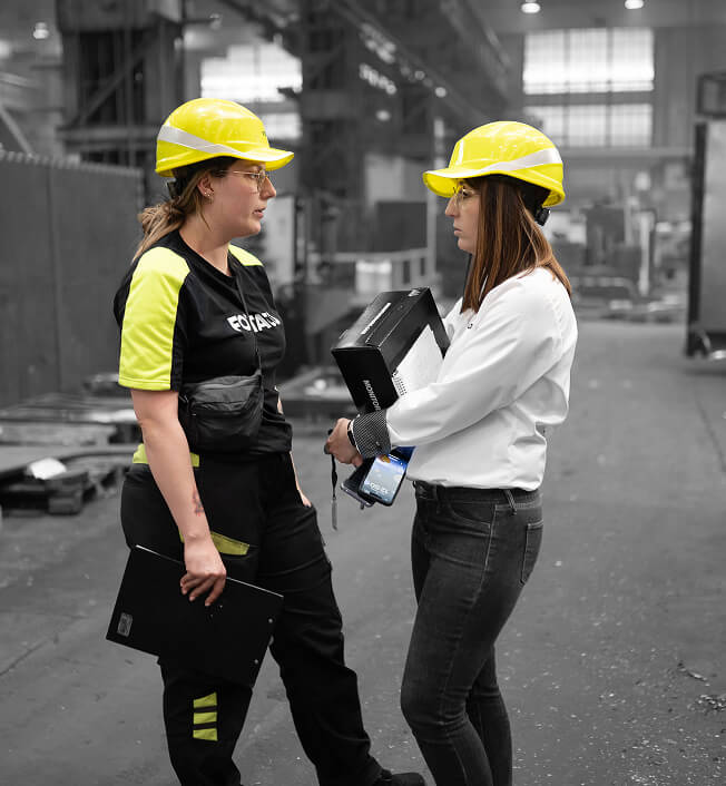 Two women wearing yellow safety helmets and holding folders have a discussion in an industrial warehouse. Both are standing, one in work attire and the other in office clothes, with machinery in the background.