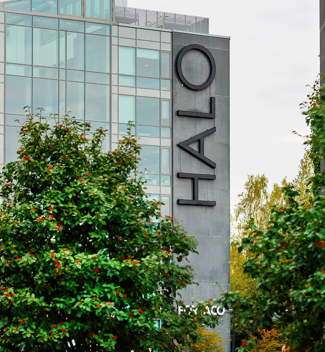 A modern glass building with the word "HALO" displayed vertically on its exterior wall, partially obscured by leafy green trees in the foreground.
