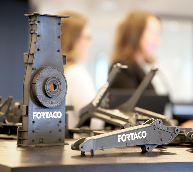 A close-up of two black metal machine parts labeled "FORTACO" on a table, with two blurred people working in the background.