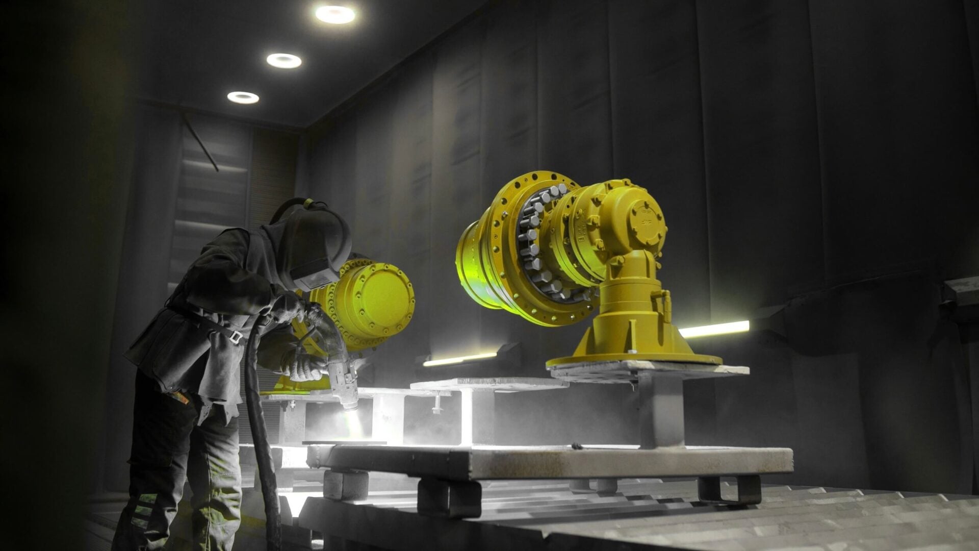 A worker in protective gear sandblasts or paints large yellow industrial machine parts inside a dimly lit workshop with bright overhead lights.