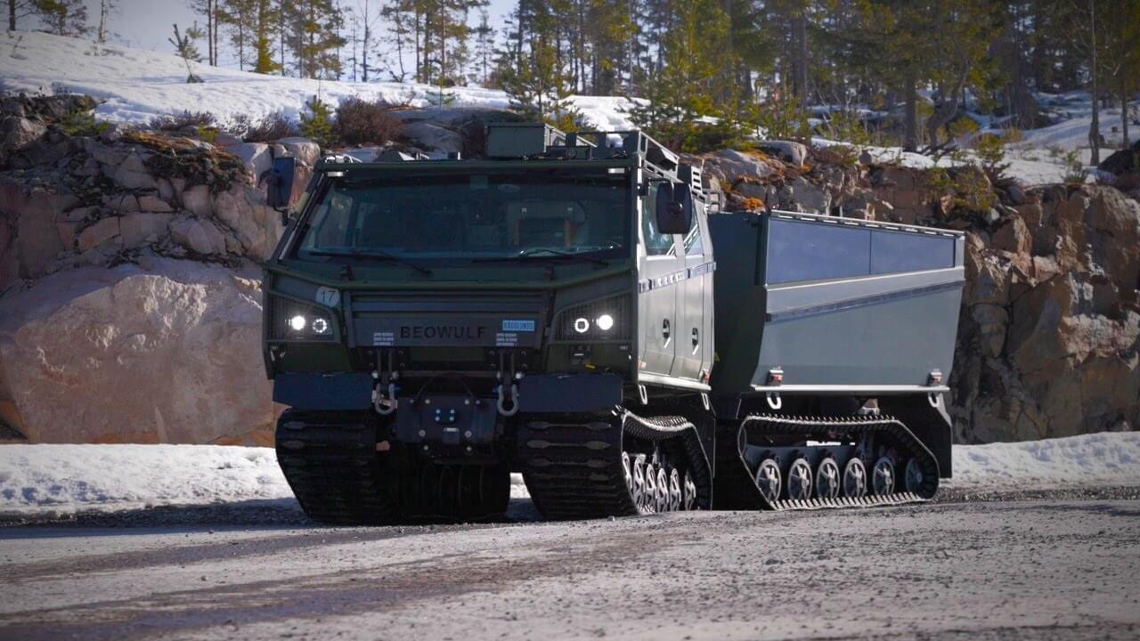 A green, tracked, articulated all-terrain vehicle labeled "Beowulf" is parked on a snowy, rocky road with pine trees and snow-covered ground in the background.