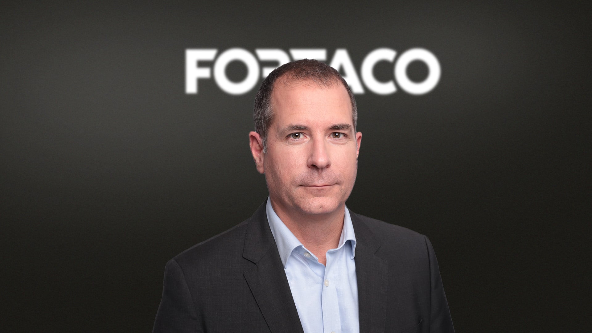 A man in a suit stands in front of a dark background with the word “FORTACO” in bold white letters behind him.