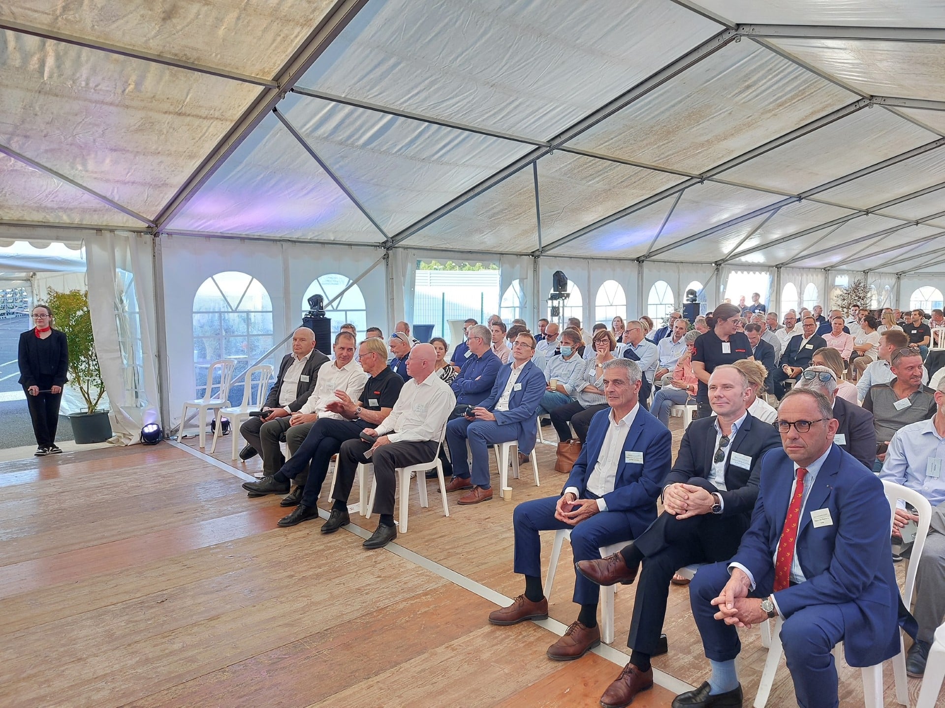 A speaker stands on the left in a large tent addressing a seated audience of professionals.
